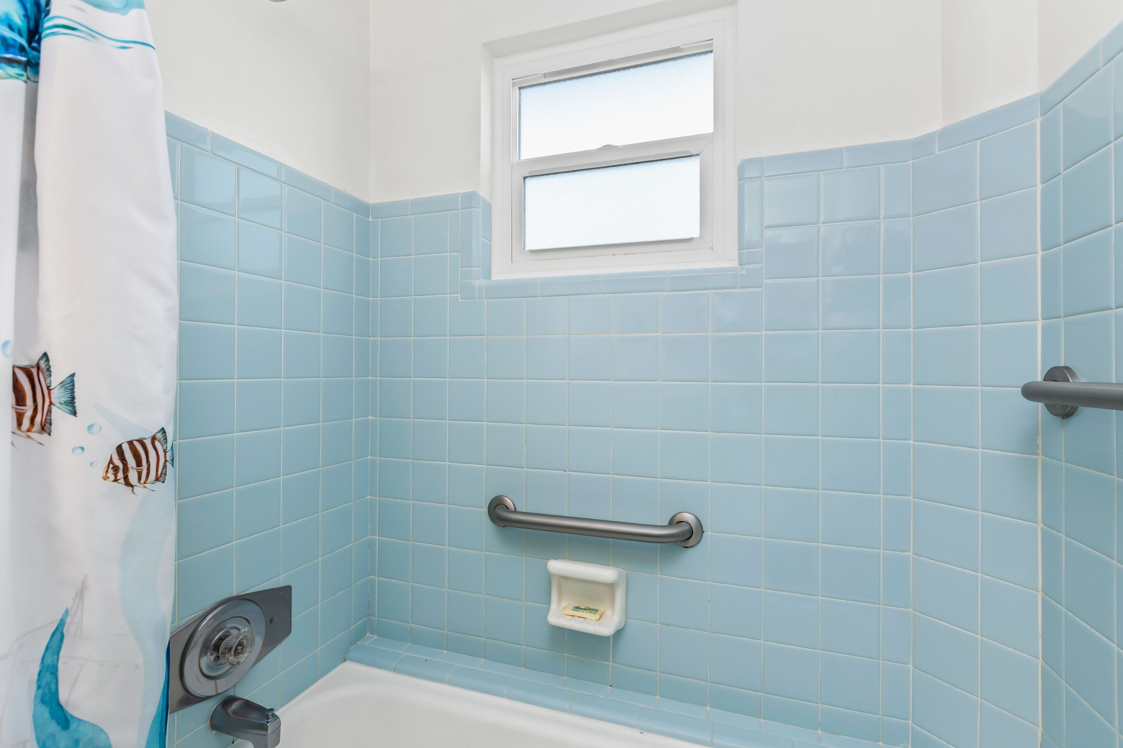 A bathroom with blue tiles, a window, a shower curtain with fish designs, a grab bar, and a soap dish.