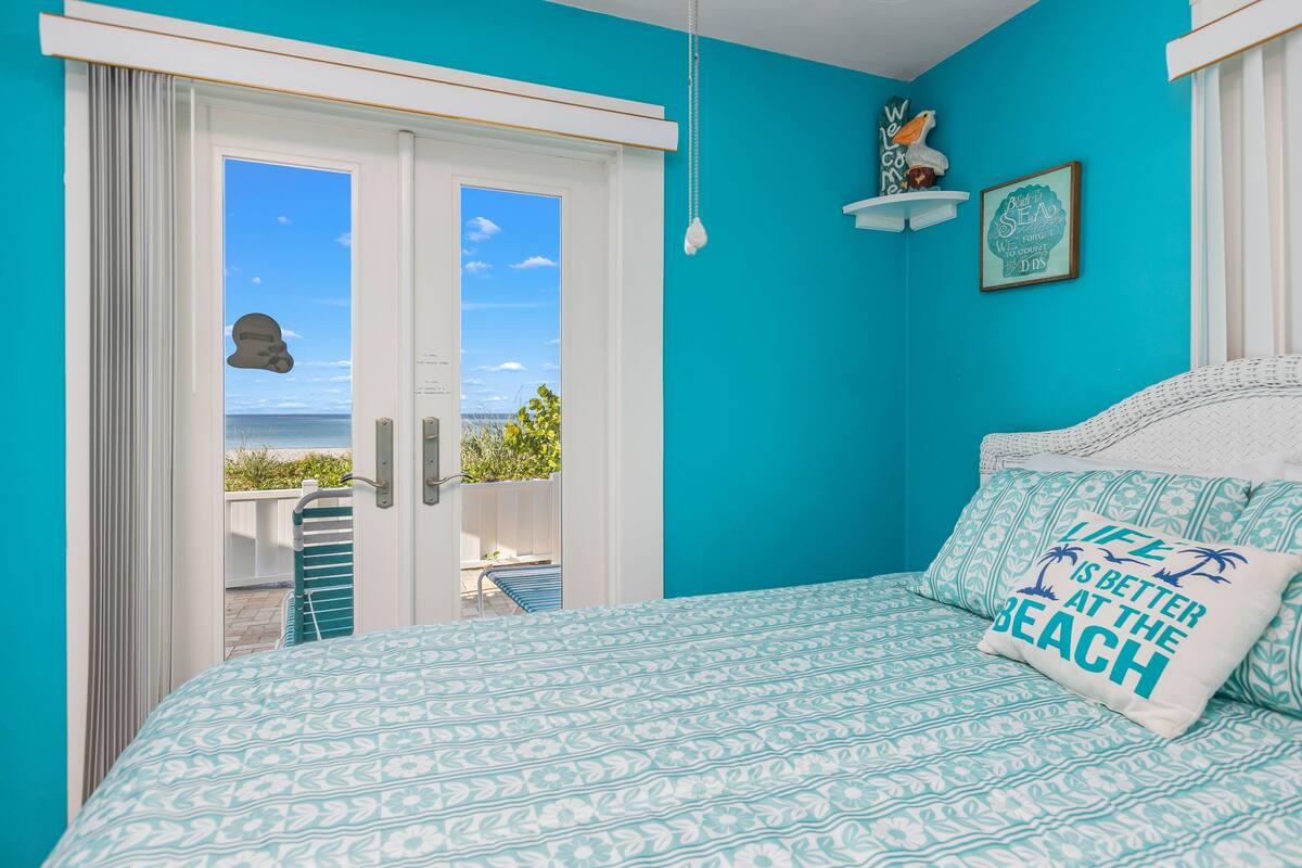Ein Schlafzimmer mit Bett, Meerblick durch eine Schiebetür und einem Kissen mit Strandmotiv.