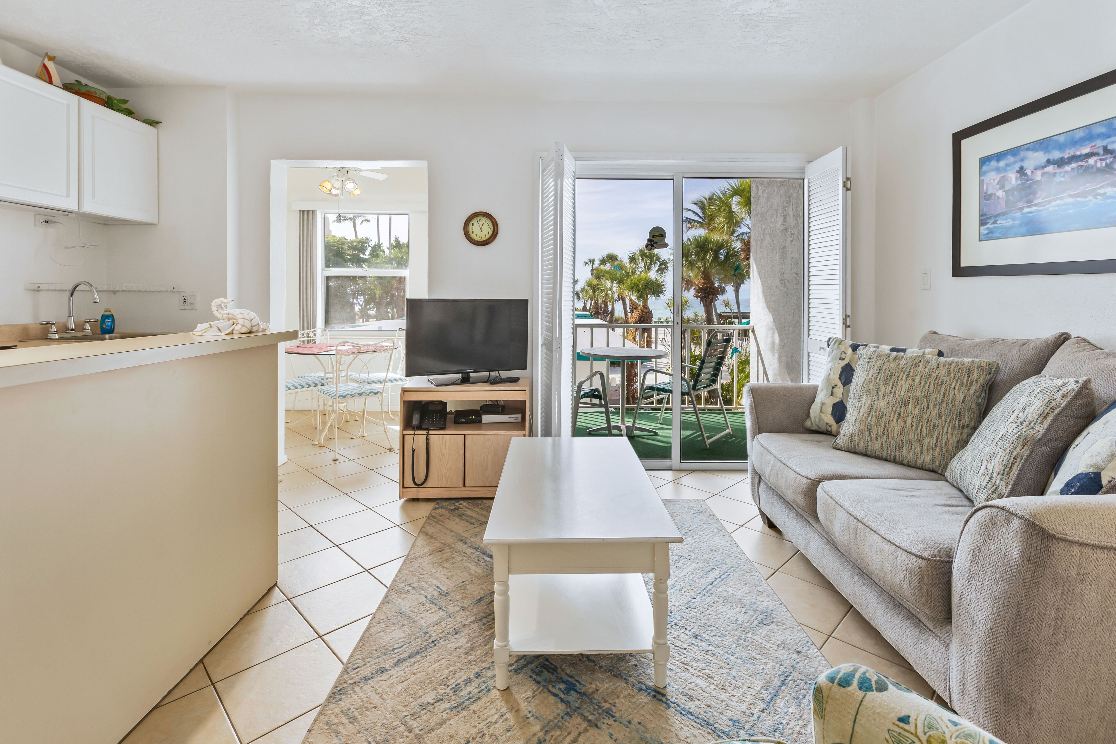 A modern living room with a sofa, a coffee table, and a TV. There is a balcony with a view of palm trees and a pool.