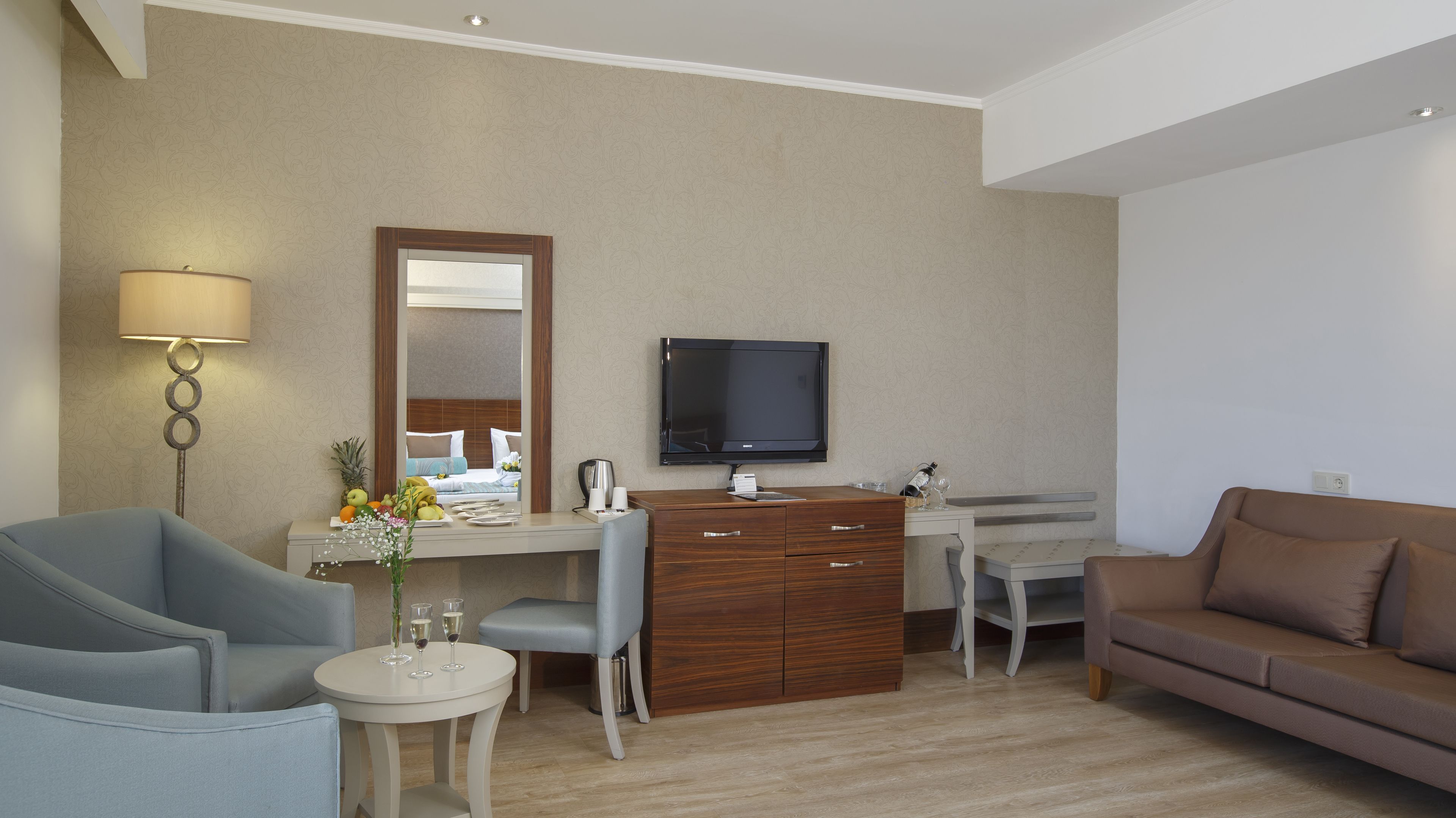 A hotel room with a bed, a desk with a flat-screen TV, a sofa, a chair, a small table with a vase and glasses, and a lamp.