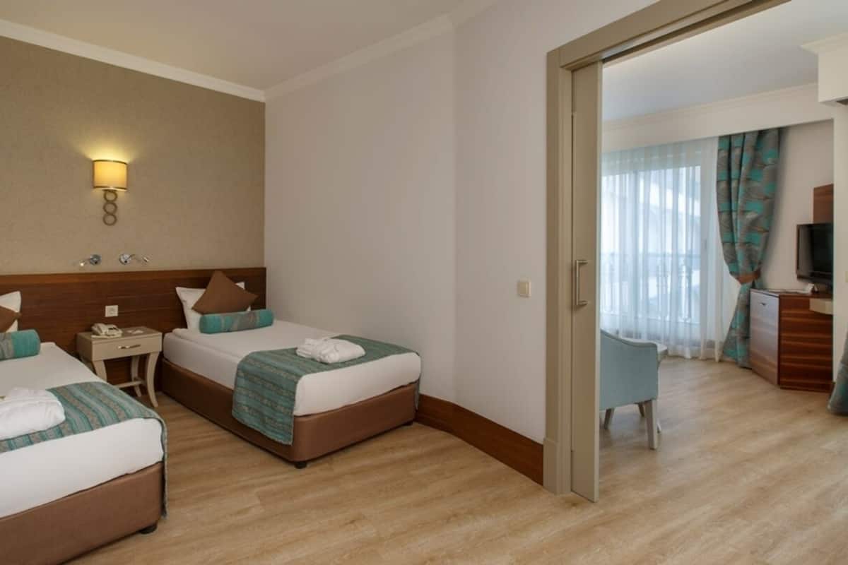 A hotel room with two beds, a bedside table with a lamp, a TV, and a view of a balcony with curtains.