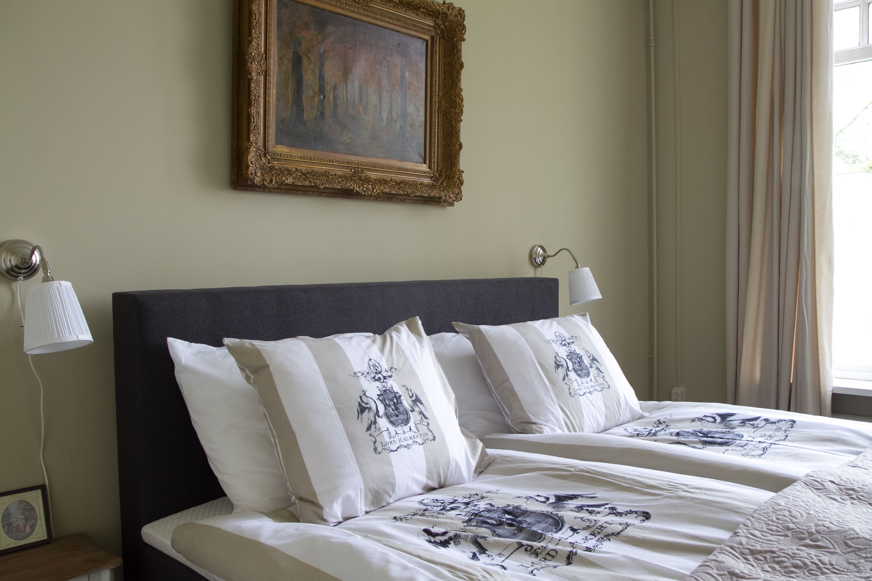 A hotel room with a large bed, two bedside lamps, a framed painting on the wall, and a window with curtains.