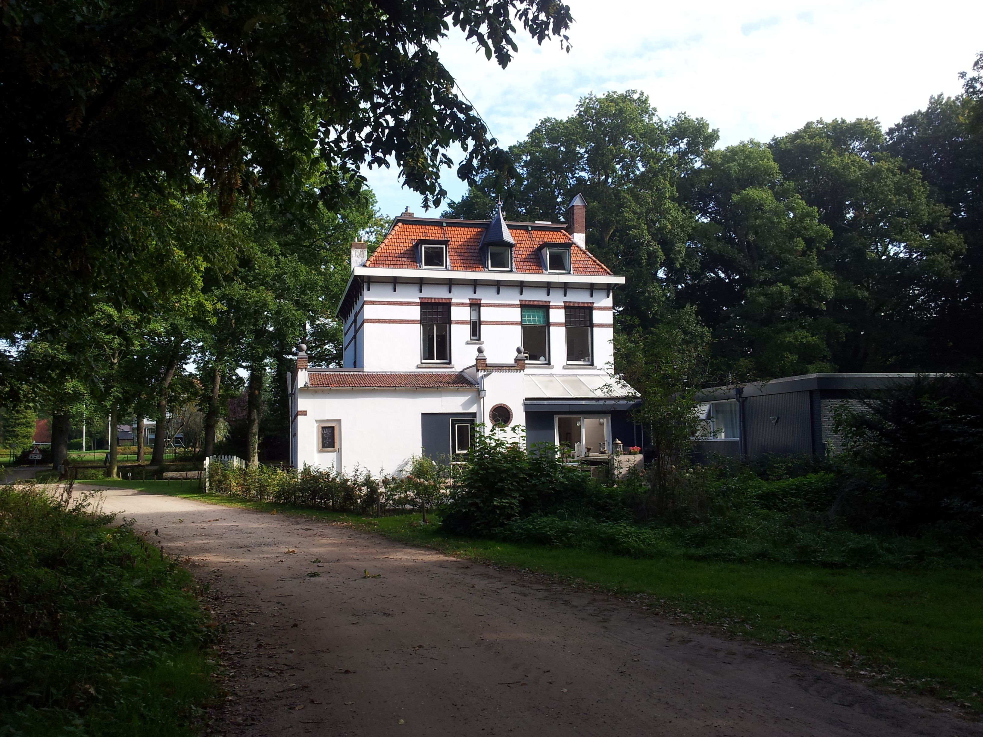 Une maison à deux étages, avec un toit recouvert de tuiles rouges, entourée d’une végétation luxuriante et un chemin de terre menant à l’entrée.