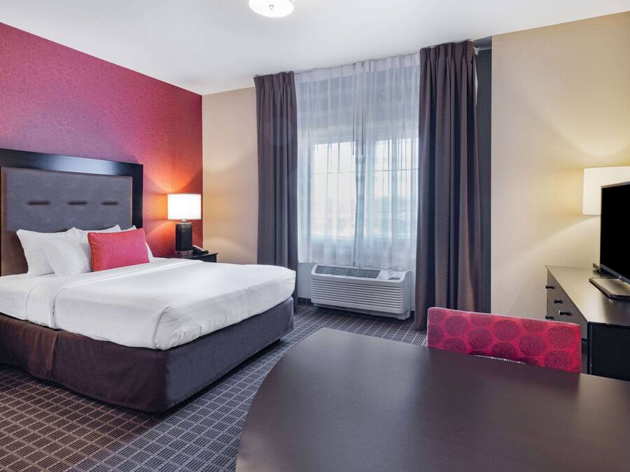 A hotel room with a bed, bedside tables, a desk, a chair, an air conditioning unit, and a television.
