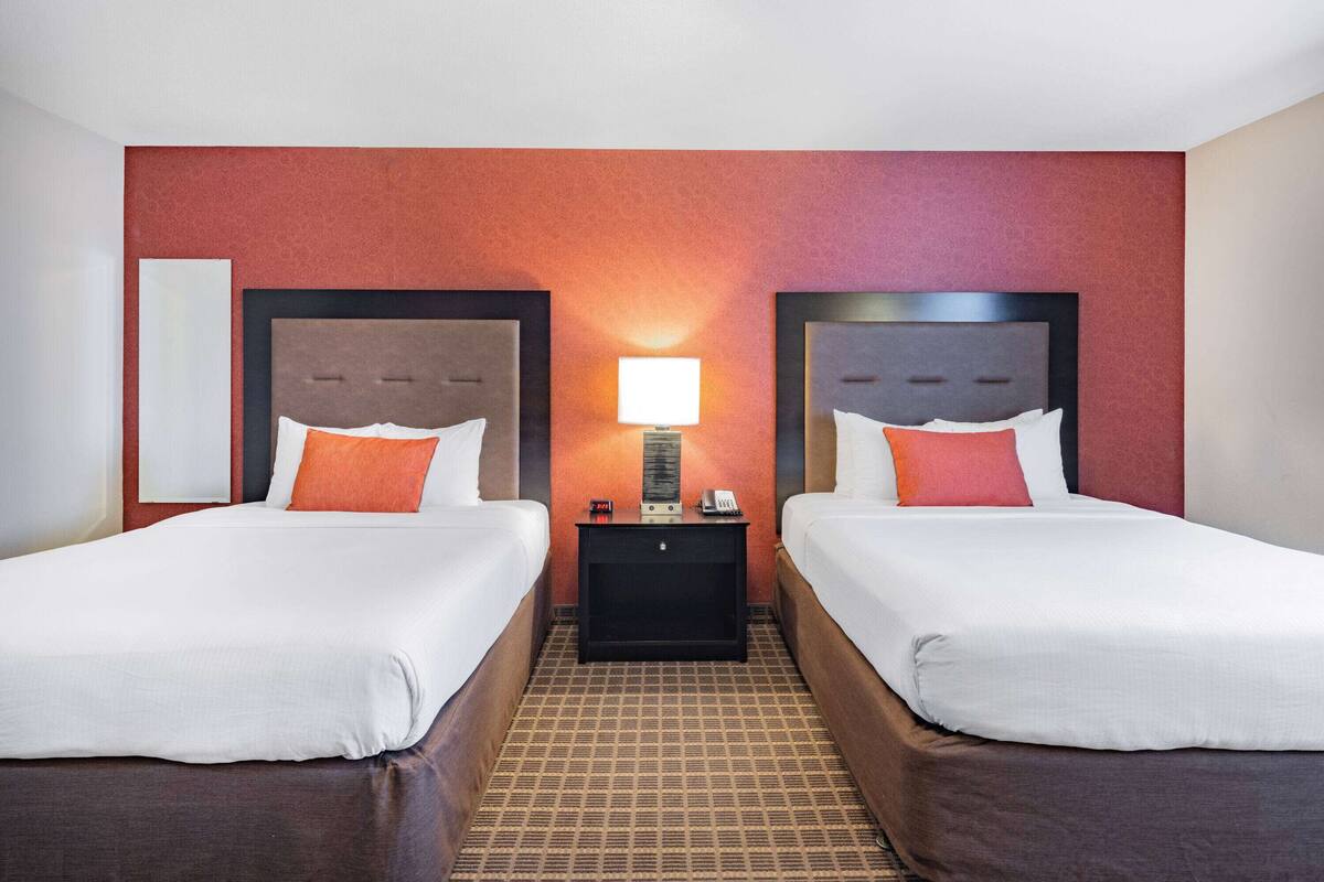 Una habitación de hotel con dos camas, una mesita de noche con lámpara y una pared roja con textura.
