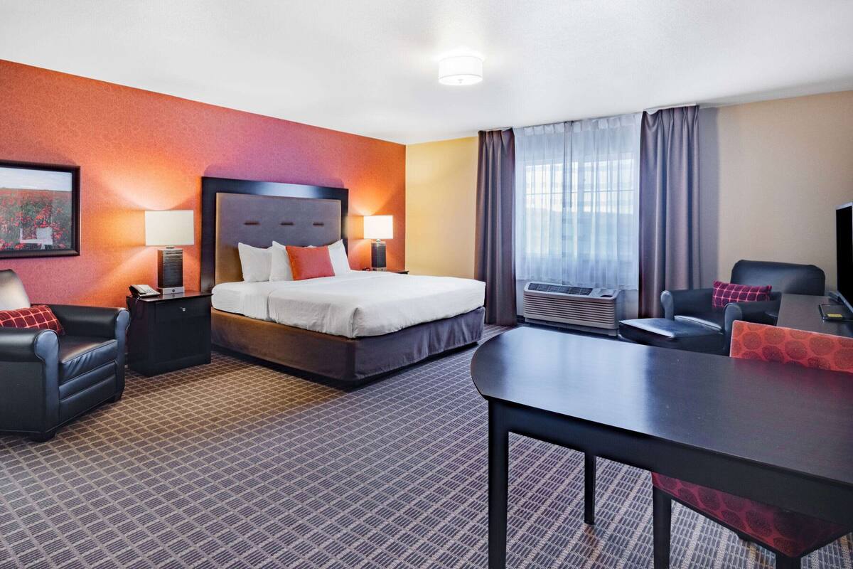 Habitación de hotel con una cama grande, dos mesitas de noche con lámparas, un escritorio con silla, un sofá y un ventanal con cortinas.