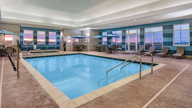 Una amplia piscina interior con agua cristalina, rodeada de tumbonas y sombrillas, y grandes ventanales que ofrecen vistas al atardecer.