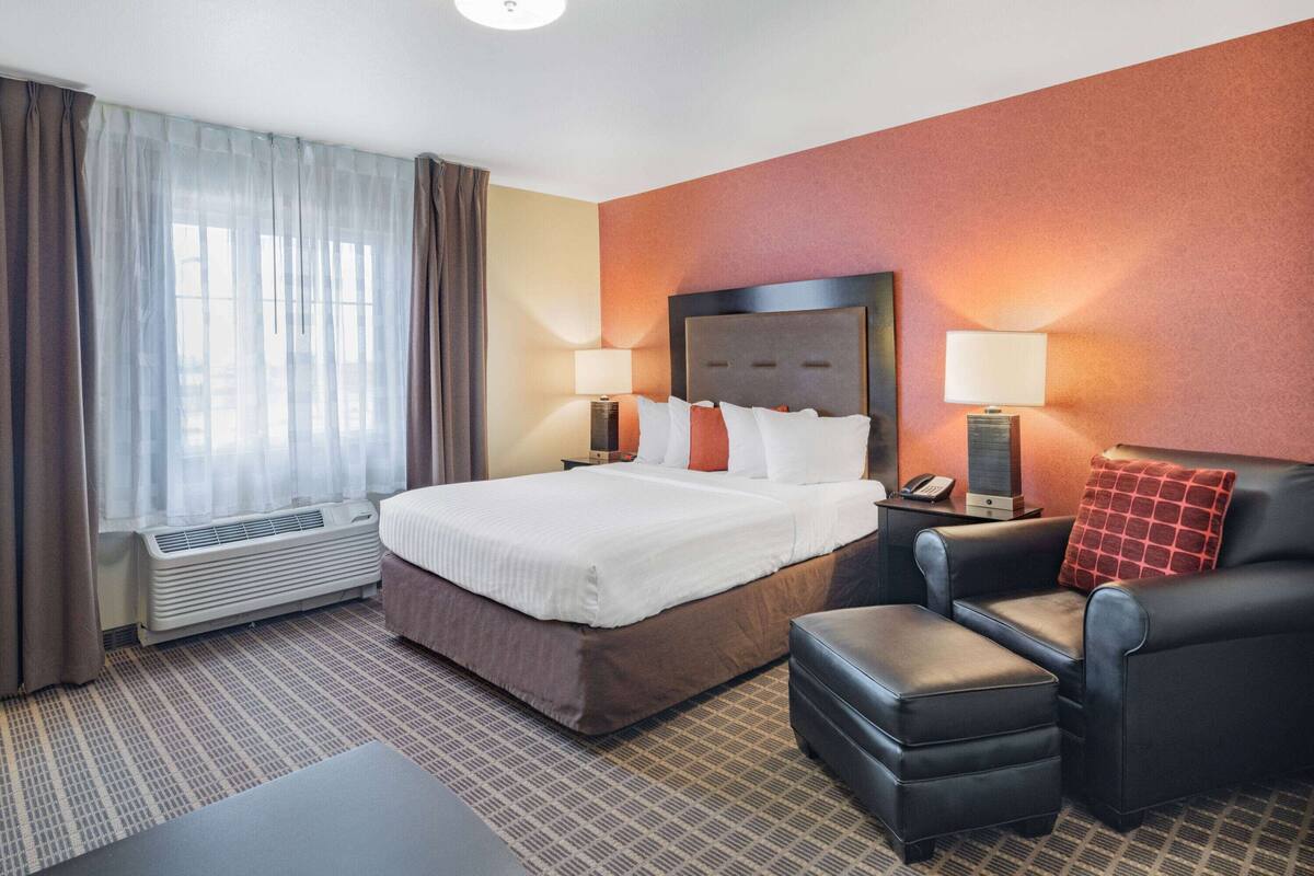 Habitación de hotel con una cama grande, un escritorio, una silla y un sofá. Se ve una ventana con cortinas, una lámpara y un calefactor.