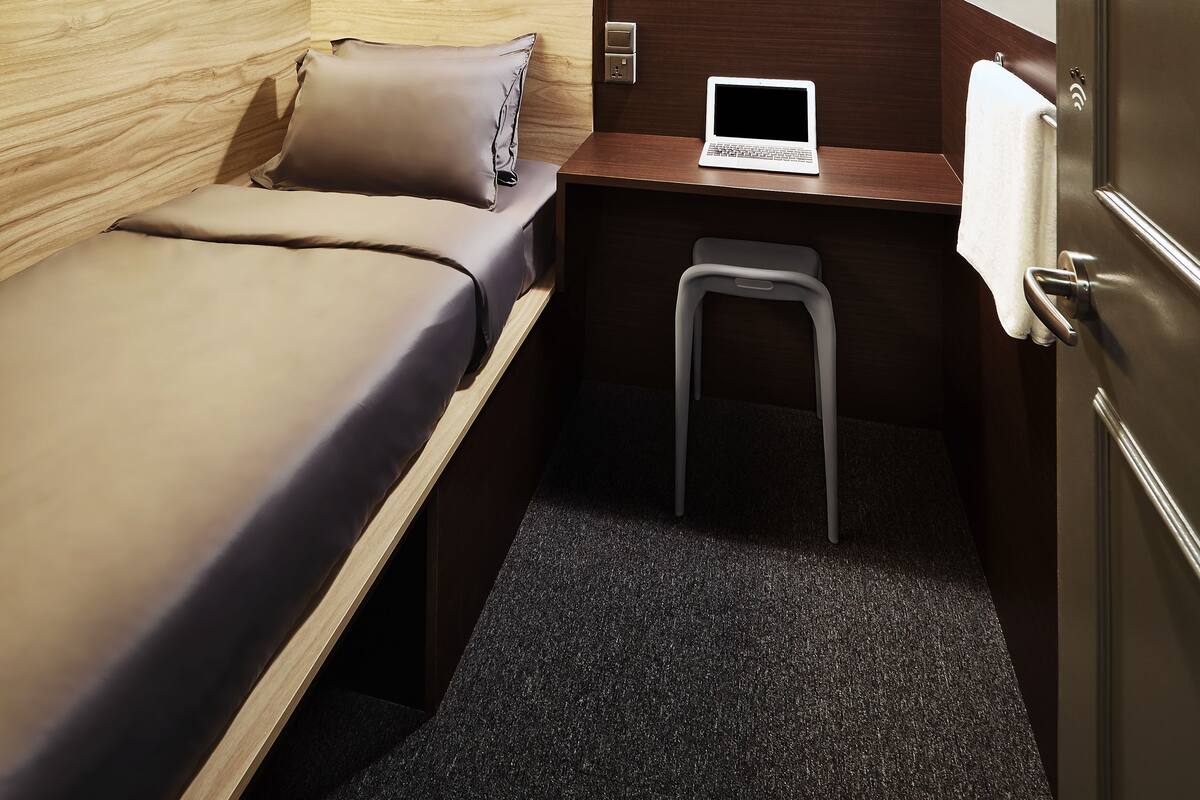 A compact room with a bed, a desk with a laptop, a small stool, and a hanging light fixture.