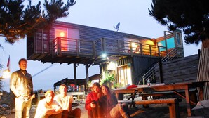 Een groep mensen verzameld rond een kampvuur in een open setting, met een houten terras en een gebouw op de achtergrond.