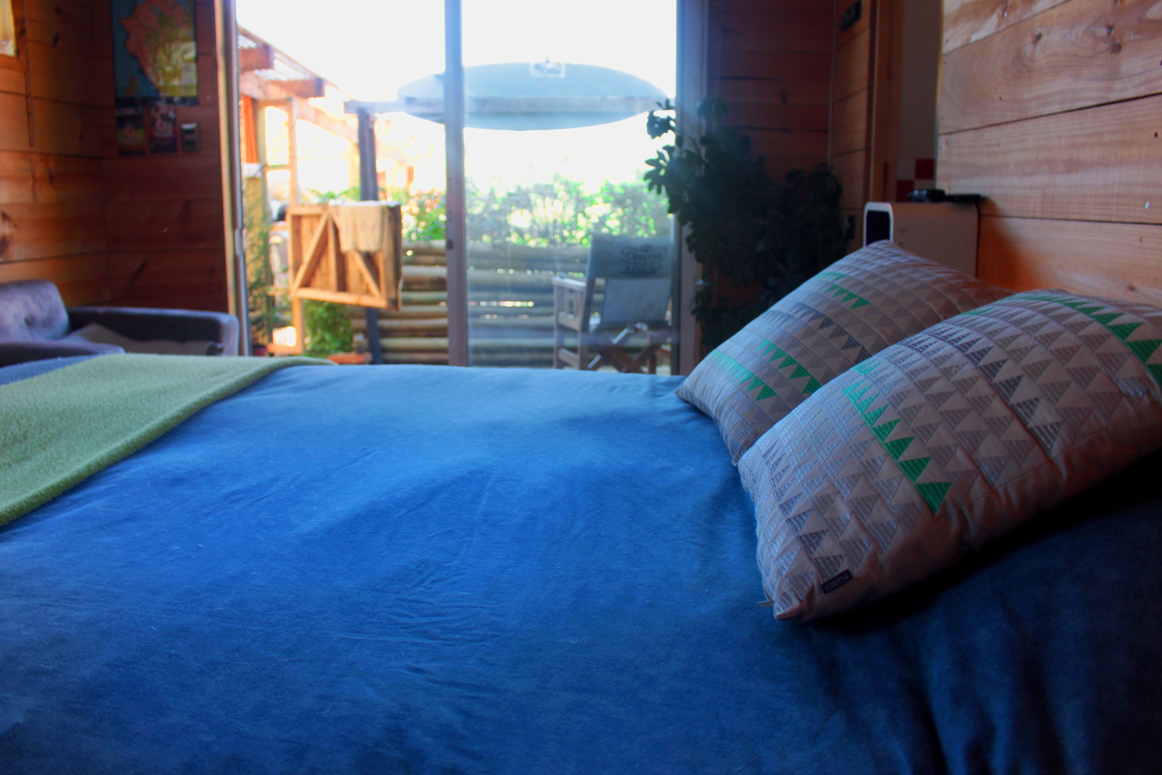 A bed with a blue mattress and a patterned pillow, a wooden chair, and a table with an umbrella outside a wooden room.