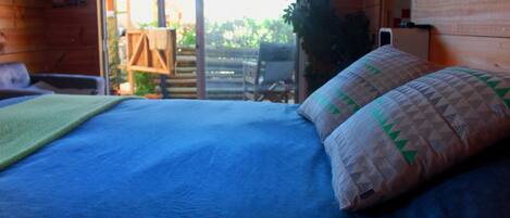 A bed with a blue mattress and a patterned pillow, a wooden chair, and a table with an umbrella outside a wooden room.