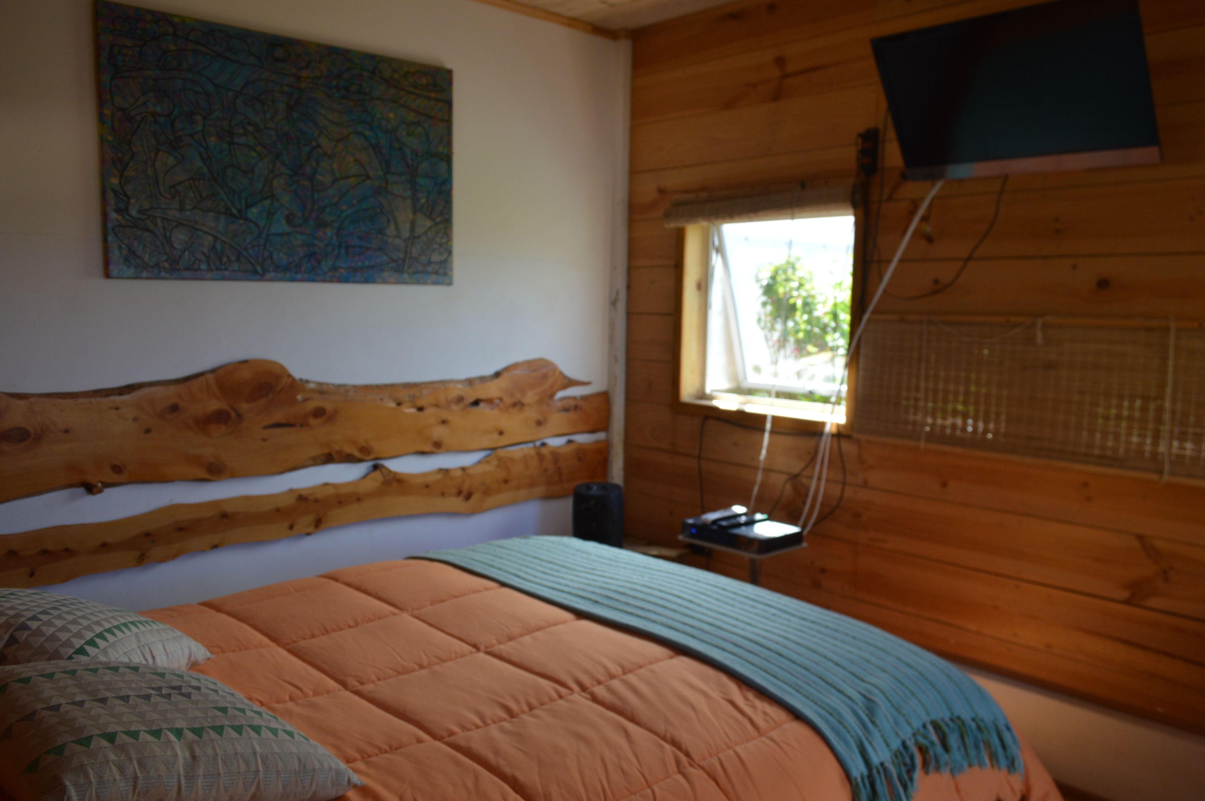 Un dormitorio con cabecera de madera, cama con colcha naranja, manta azul, un mapa en la pared y una ventana con vistas a zonas verdes.