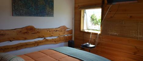 Un dormitorio con cabecera de madera, cama con colcha naranja, manta azul, un mapa en la pared y una ventana con vistas a zonas verdes.