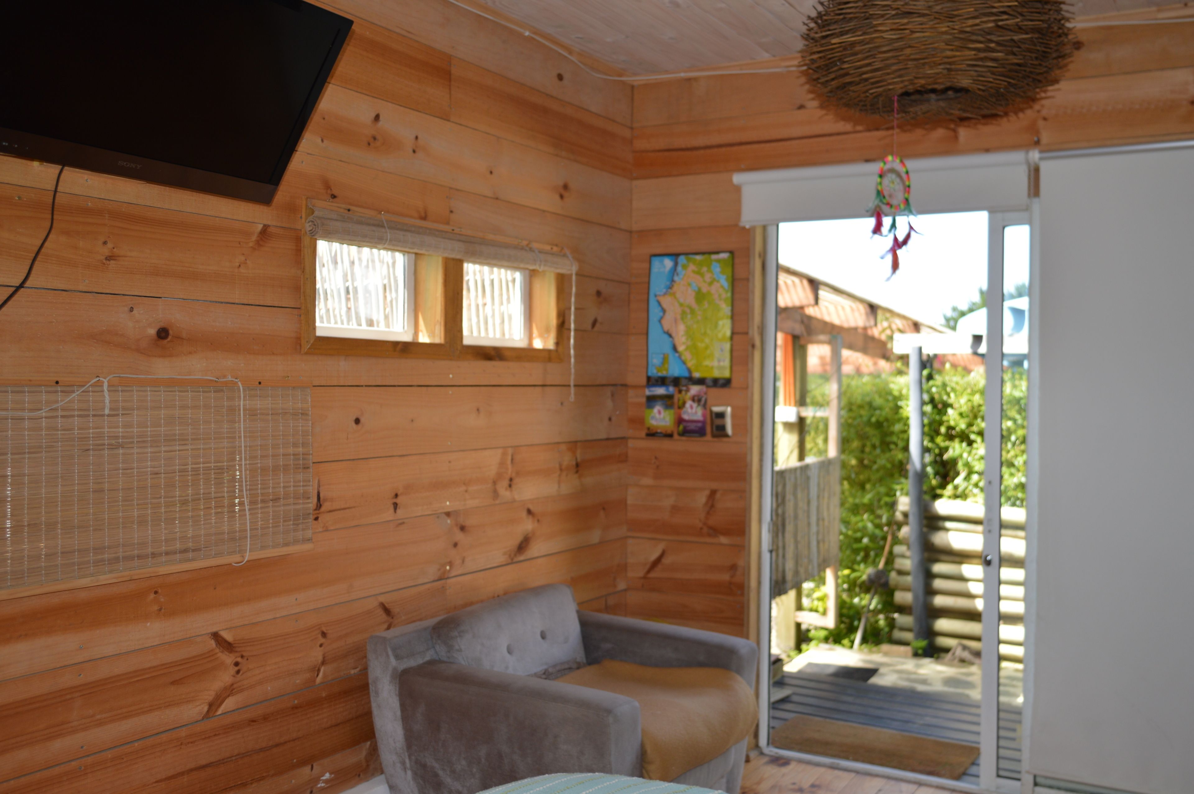 A wooden cabin interior with a sofa, a television, a window with blinds, and a door leading outside.