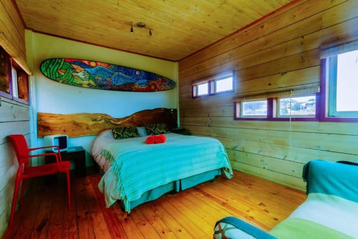 Un dormitorio de cabaña de madera con una cama, una silla roja, una ventana y una obra de arte colorida en la pared.