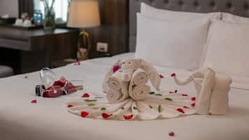 A neatly made bed with a towel arrangement and rose petals, a bedside table with a lamp, and a window with flowers.