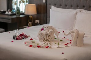 Cama bem arrumada com arranjo de toalhas e pétalas de rosa, mesa de cabeceira com abajur e janela com flores.