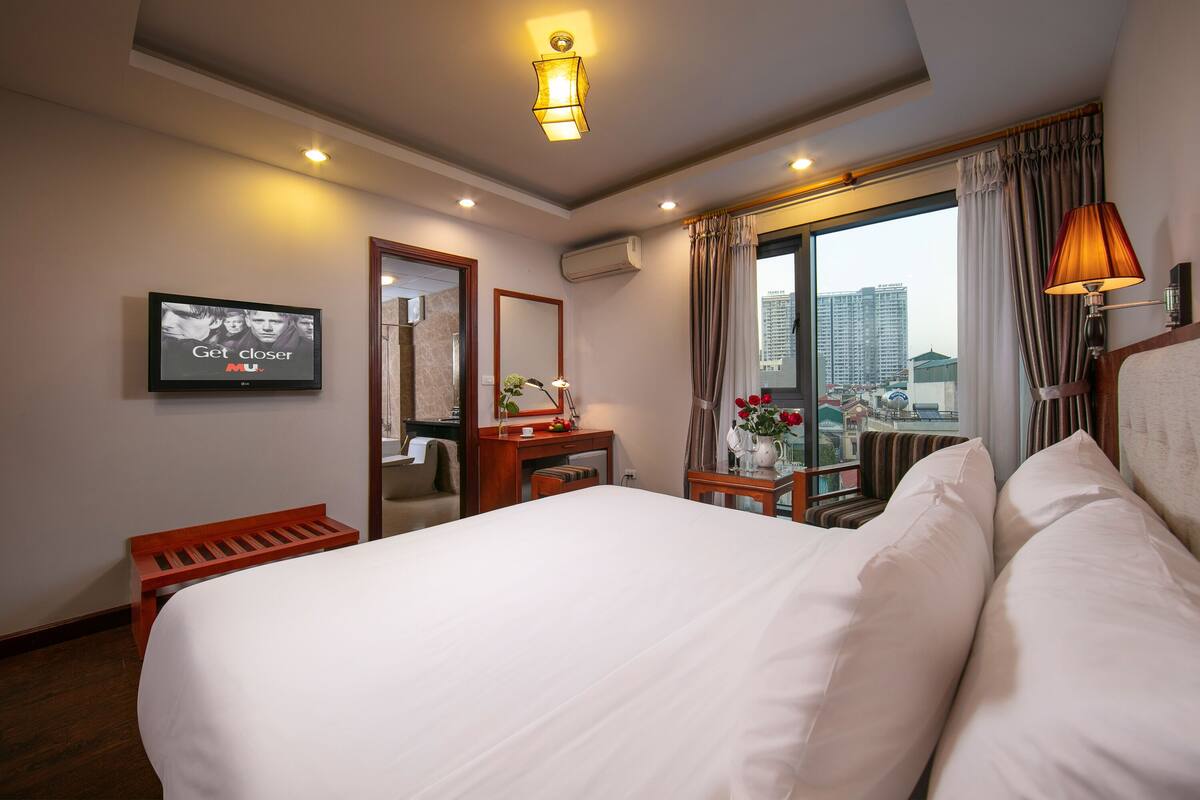 A hotel room with a large bed, a desk with a chair, a small table with a vase of flowers, a television on the wall, and a window with curtains.