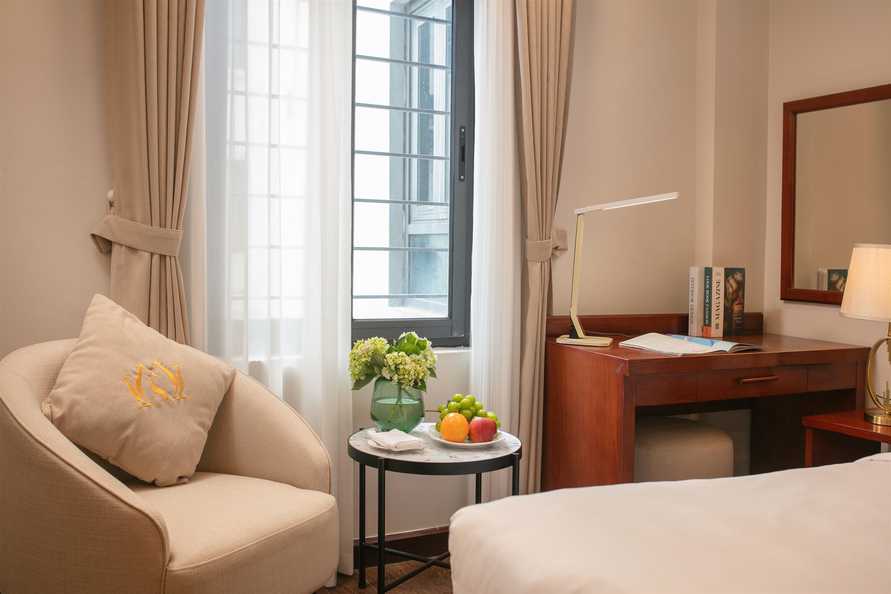 Habitación de hotel con cama, escritorio, silla, una pequeña mesa con un florero y fruta, y ventana con cortinas.