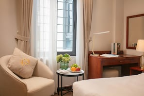 A hotel room with a bed, a desk, a chair, a small table with a vase and fruit, and a window with curtains. - Sen Luxury Hotel (Hanoi)