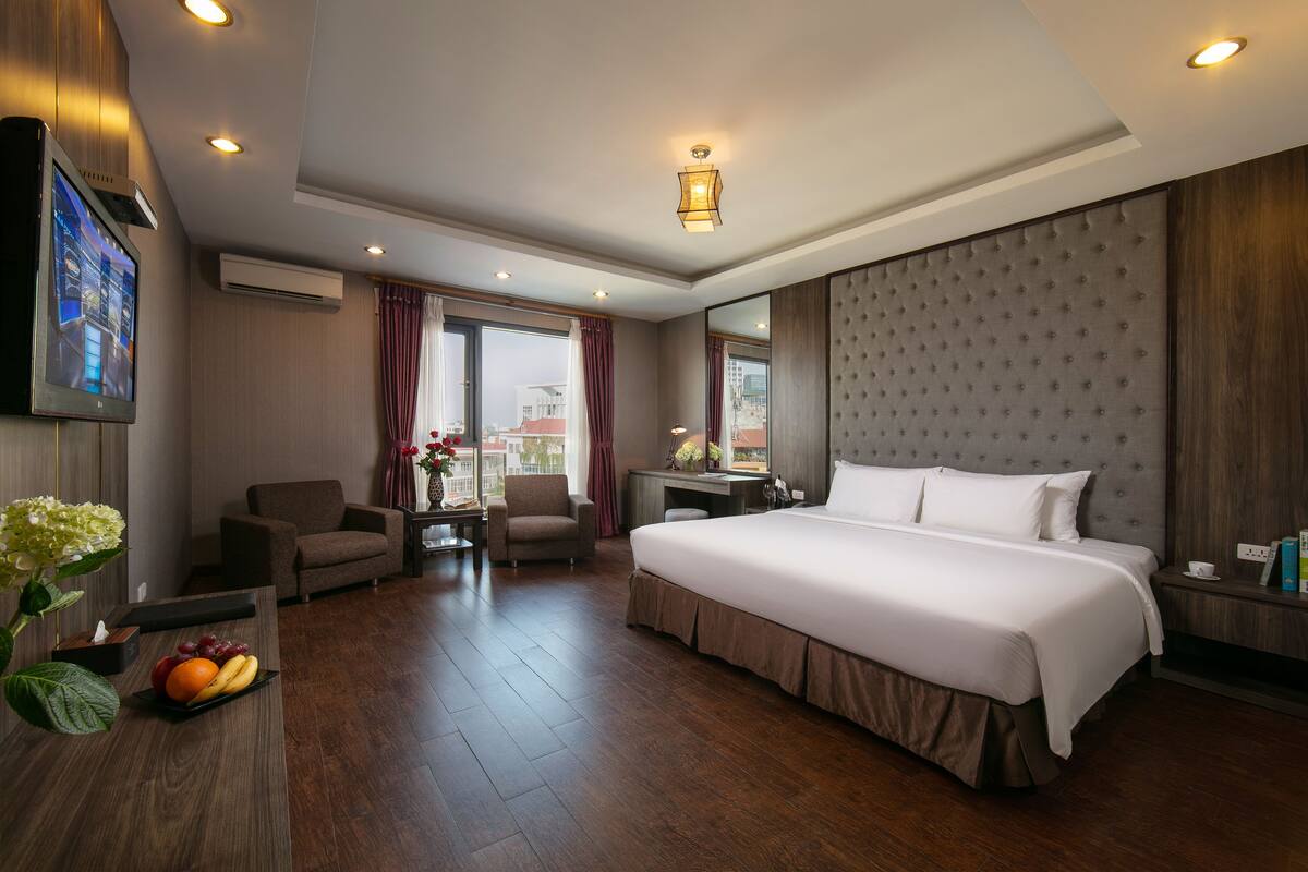 A hotel room with a large bed, a desk, two chairs, a TV, and a window with curtains.