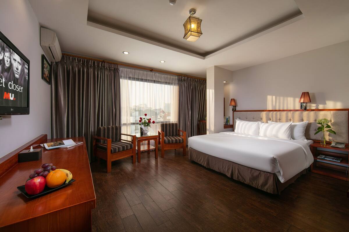 A hotel room with a large bed, a desk with a fruit tray, a chair, a TV, and a window with curtains.