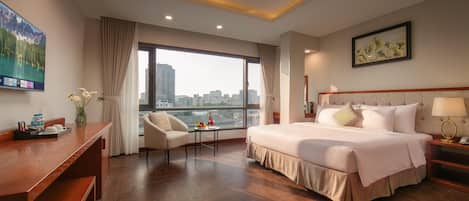 A hotel room with a bed, a desk, a chair, a window with a city view, and a painting on the wall.