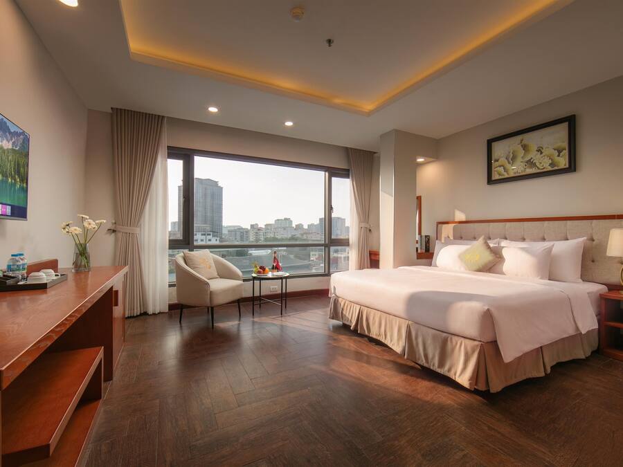 Habitación de hotel con cama, escritorio, silla, ventana con vista a la ciudad y un cuadro en la pared.
