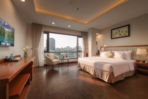 A hotel room with a bed, a desk, a chair, a window with a city view, and a painting on the wall. - Sen Luxury Hotel (Hanoi)