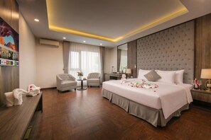 A hotel room with a large bed, two armchairs, a small table with flowers, and a large mirror.