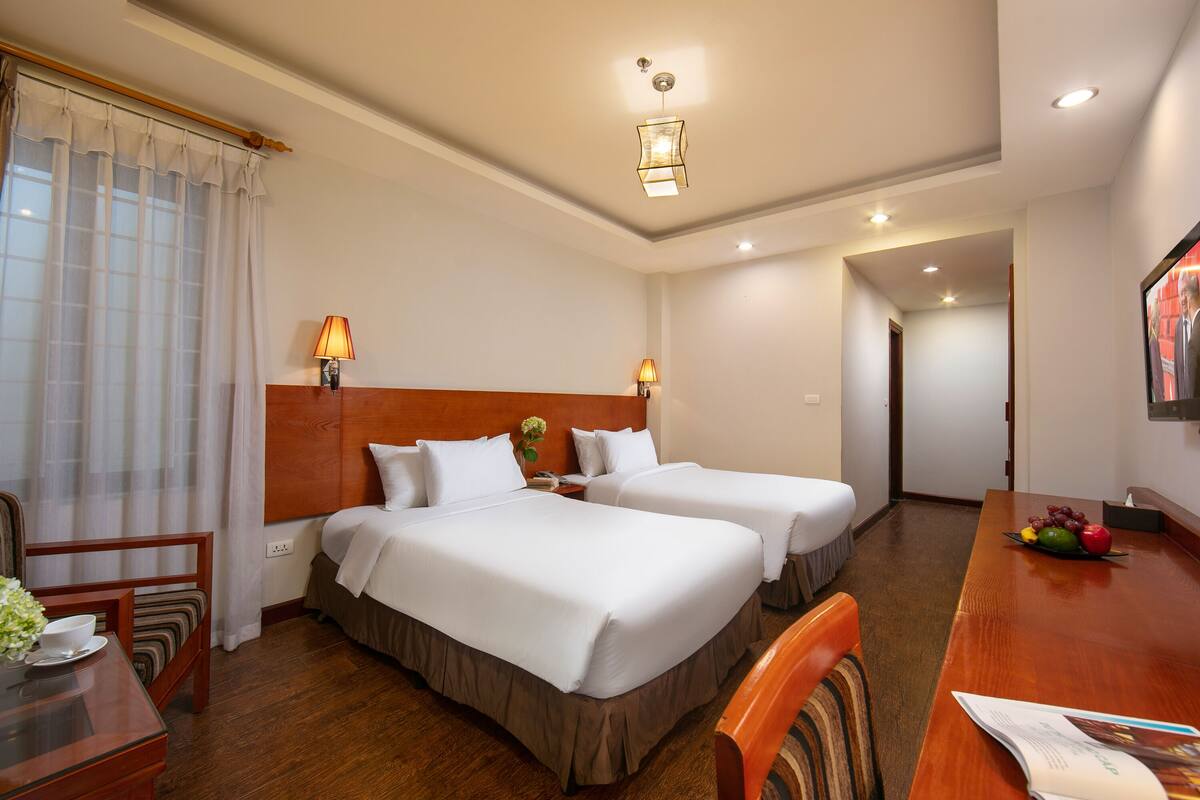 A hotel room with two beds, a wooden headboard, a dining table with a fruit bowl, a chair, and a lamp.