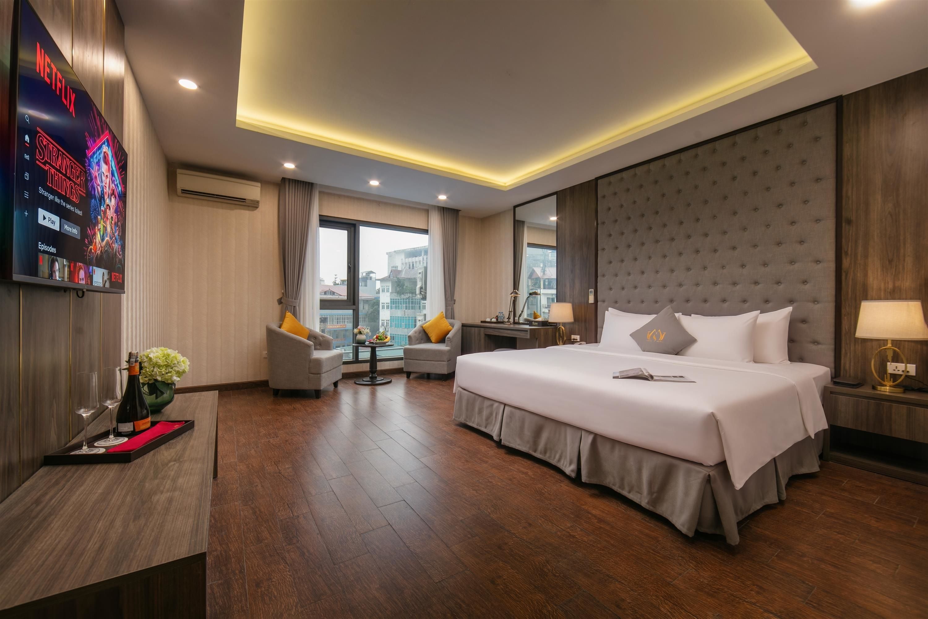 A hotel room with a large bed, a TV mounted on the wall, a desk with a chair, and a view of the city through the window.