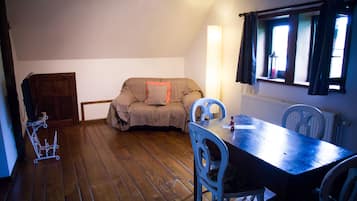 Una habitación con suelo de madera, un sofá con cojín, una mesa con sillas, un radiador y una ventana con cortinas.