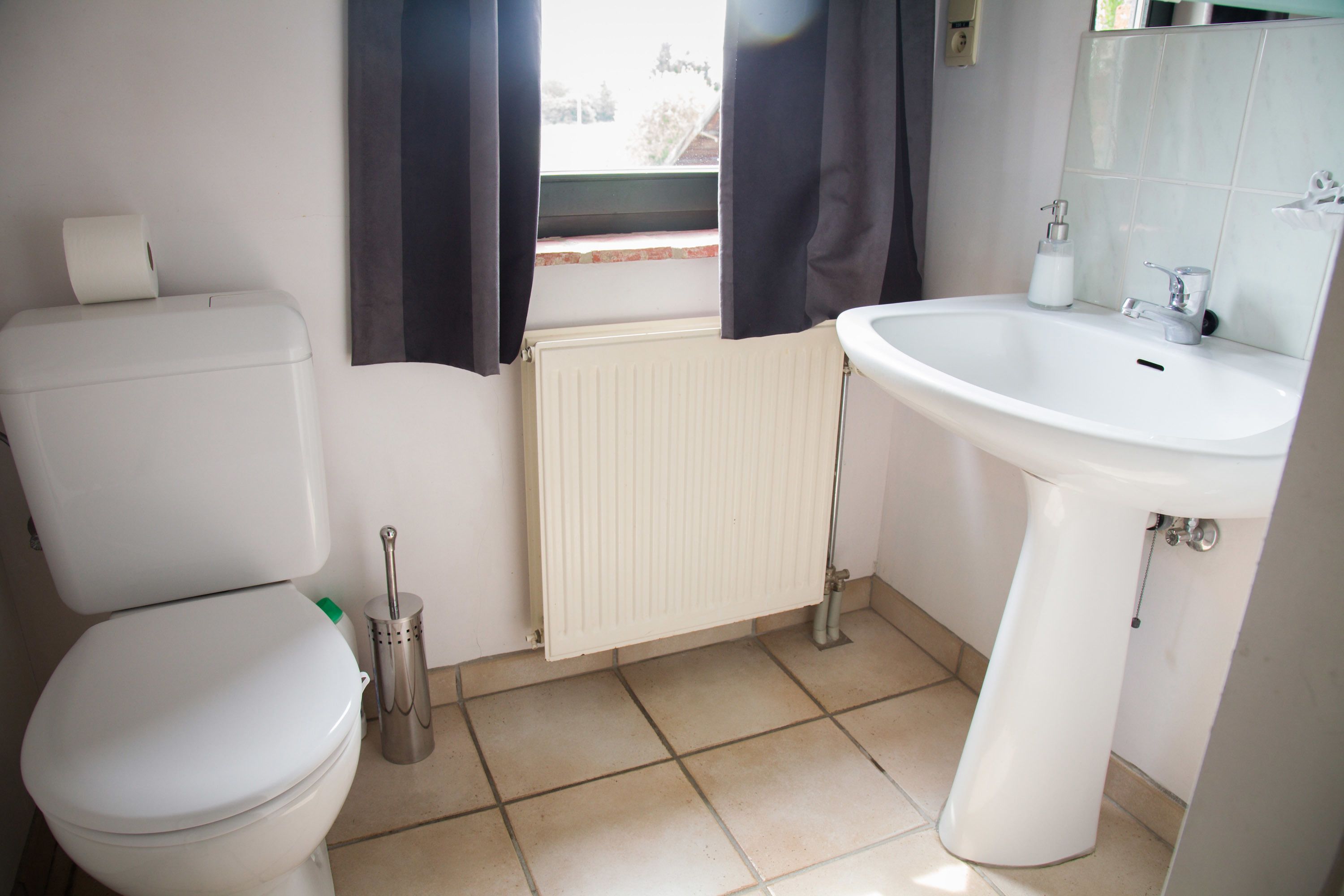 A bathroom with a toilet, a radiator, a sink, and a window with curtains.