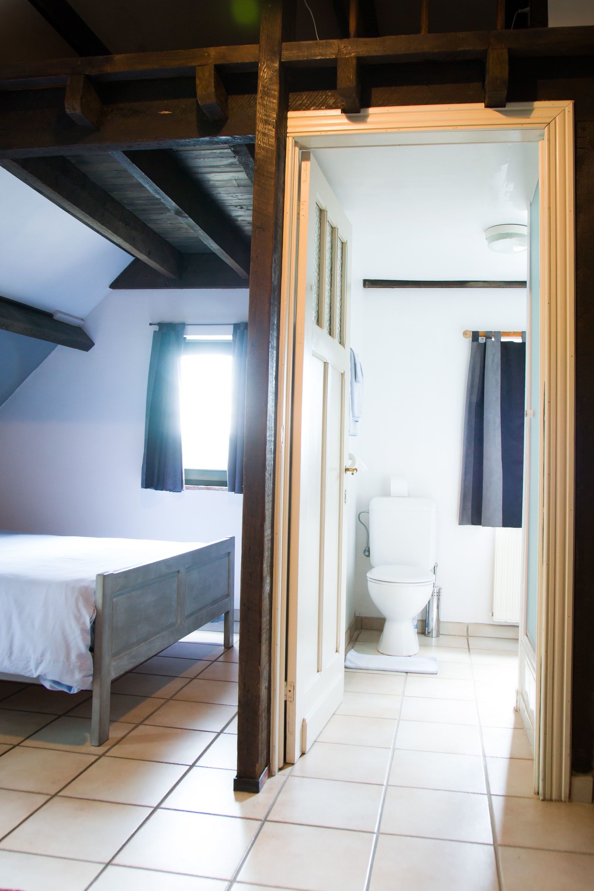 A room with a bed, a window with curtains, a white toilet, and a door leading to another room.