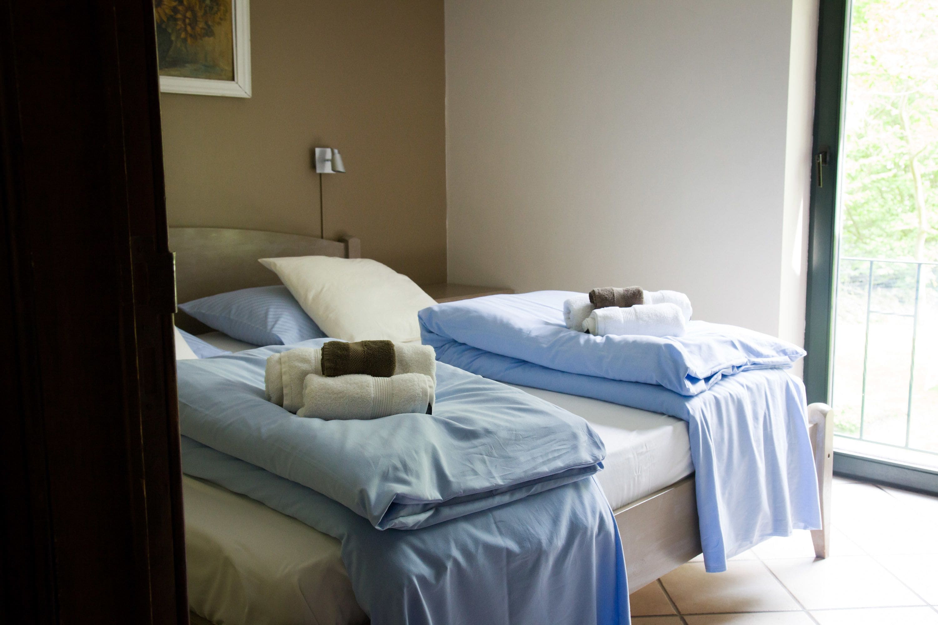 A hotel room with two beds, each with blue bedding and a white pillow. There is a lamp on a bedside table and a window with a view of greenery outside.