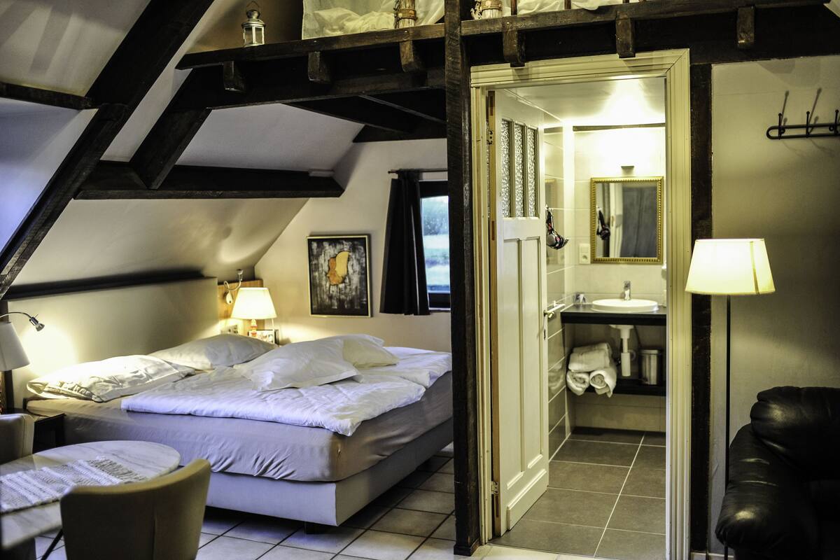Une chambre à coucher comprenant un lit, des tables de chevet, une chaise, un bureau, et une salle de bain avec un lavabo, un miroir et un porte-serviettes.