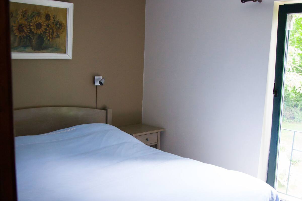 Une chambre à coucher comprenant un lit, une table de chevet, une lampe fixée au mur et une fenêtre donnant sur un paysage verdoyant.