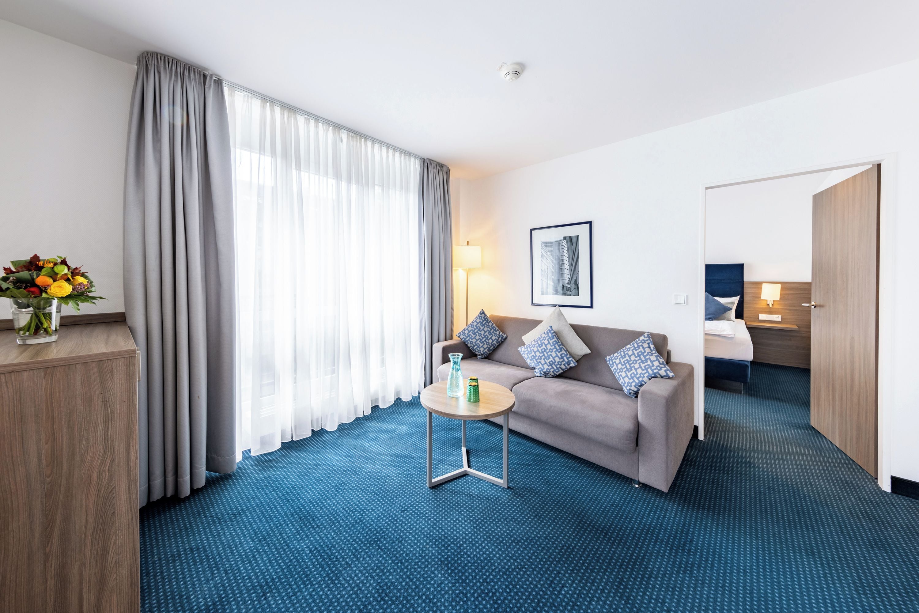 Une chambre d’hôtel avec un canapé, une petite table, une lampe, un tableau au mur et une grande fenêtre avec des rideaux.