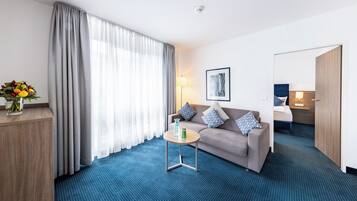 Une chambre d’hôtel avec un canapé, une petite table, une lampe, un tableau au mur et une grande fenêtre avec des rideaux.