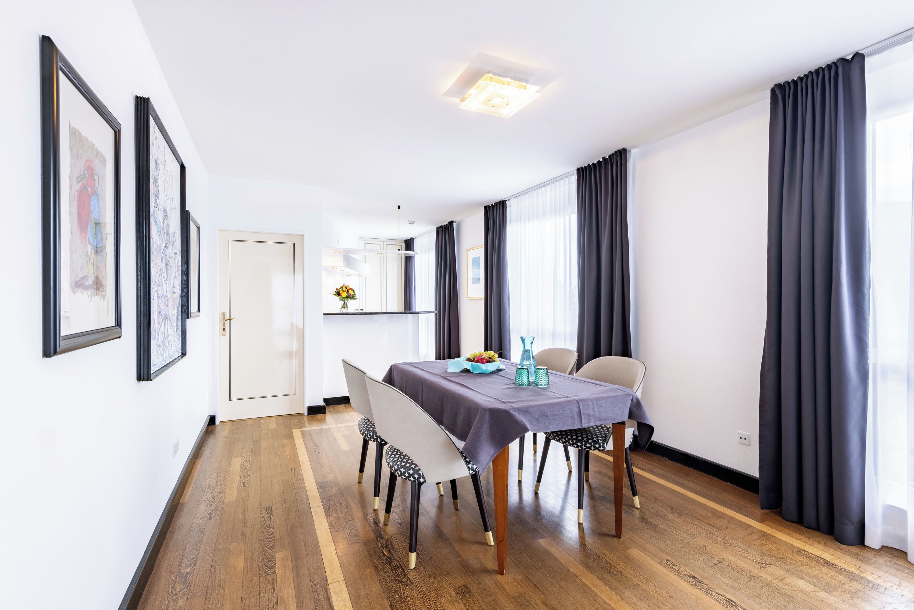 Een eetgedeelte met een tafel gedekt voor vier personen, houten vloeren, en uitzicht op een keuken en eetruimte via een deuropening.