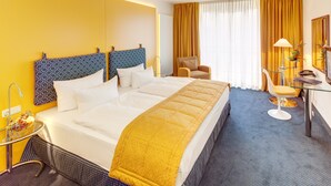 A hotel room with a large bed, a desk with a chair, a small table with a lamp, and a window with curtains. - Hotel Alexander am Zoo (Frankfurt)