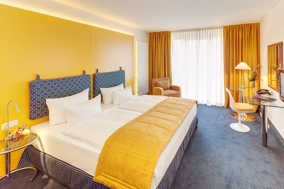 A hotel room with a large bed, a desk with a chair, a small table with a lamp, and a window with curtains.