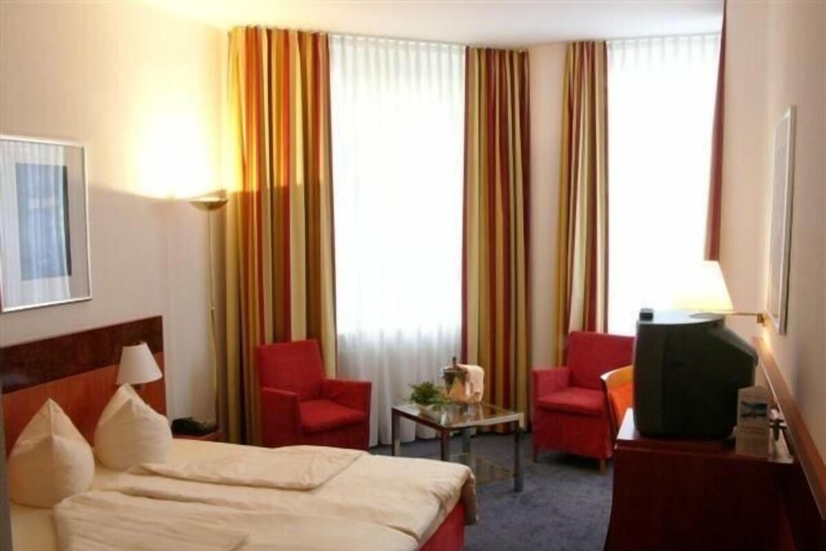 Hotellihuone, jossa on sänky, kaksi punaista tuolia, kirjoituspöytä lampun kanssa ja televisio.