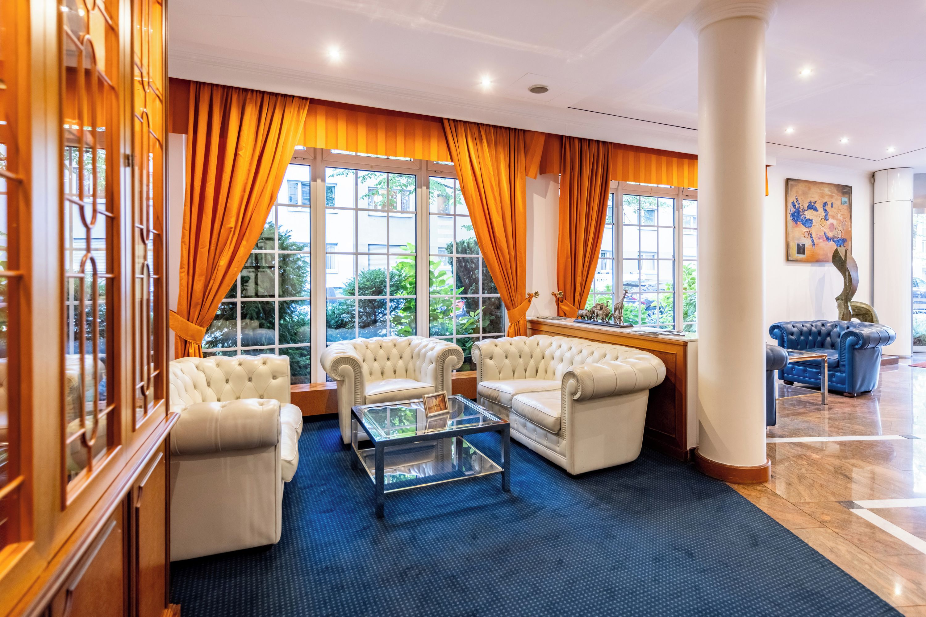 Un hall d’hôtel doté d’un espace salon comprenant deux canapés en cuir blanc, une table basse en verre et une moquette bleue.