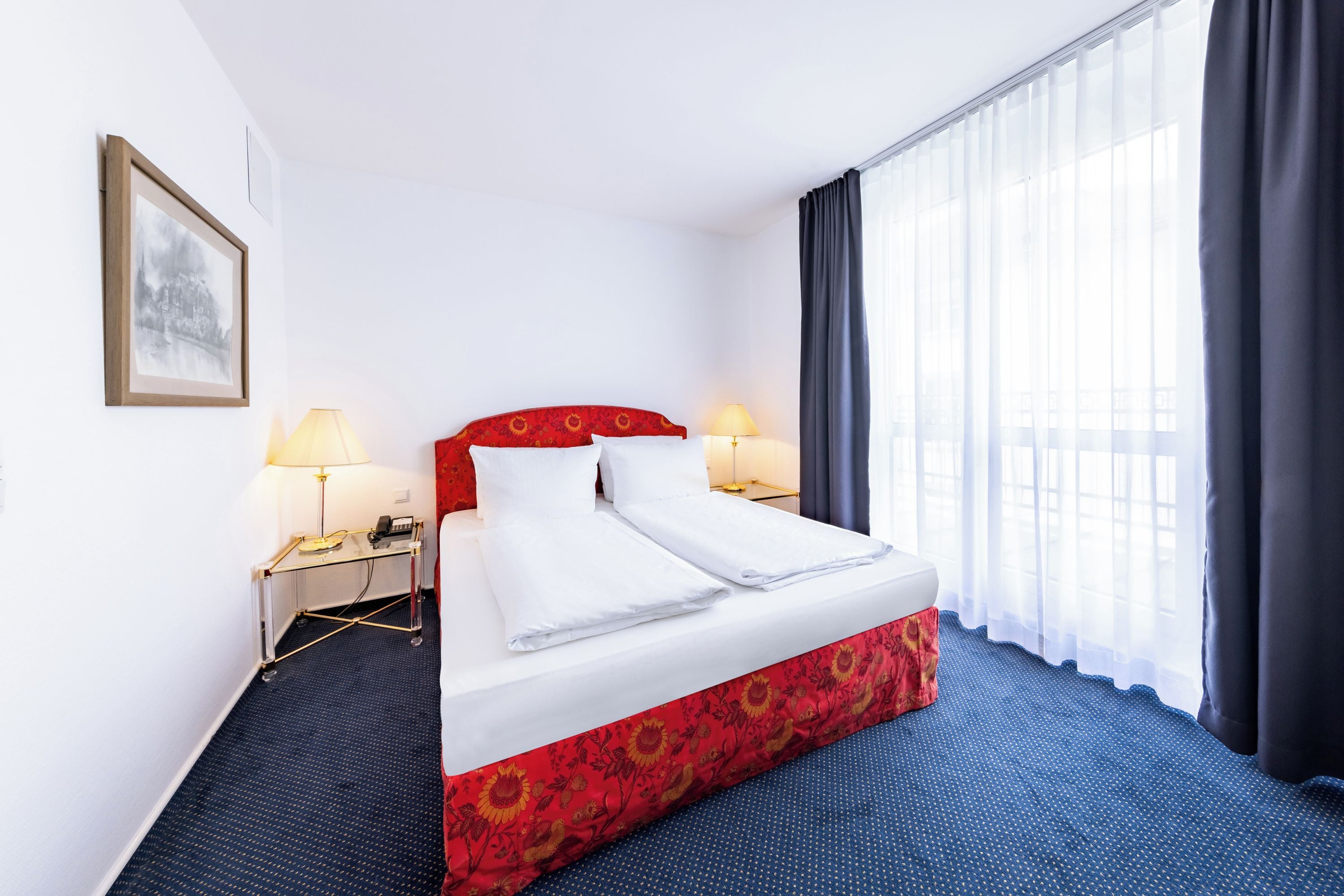 Une chambre d’hôtel avec un lit, deux lampes de chevet, un téléphone, un tableau encadré au mur et une fenêtre avec des rideaux.