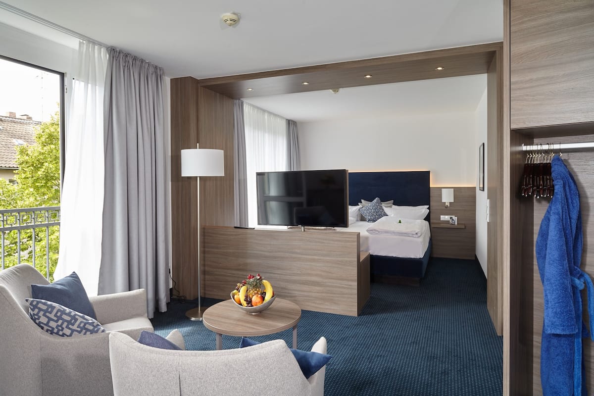 A hotel room with a bed, a TV, a desk, a chair, a small table with fruit, and a balcony with curtains.