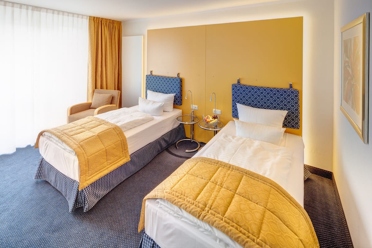 A hotel room with two beds, a small table with a fruit bowl, a chair, and a large window with curtains.