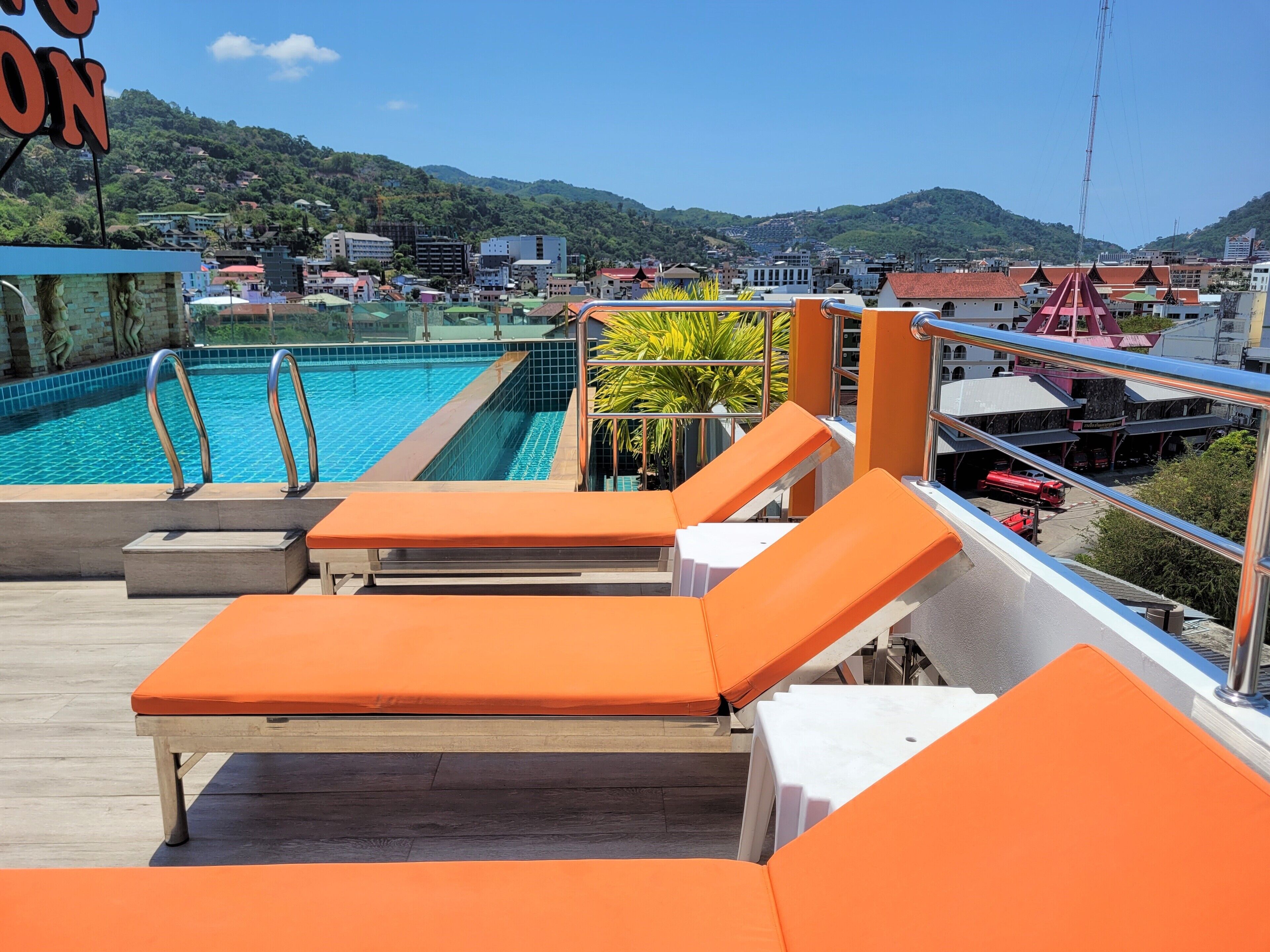A rooftop pool area with orange lounge chairs, a swimming pool, and a view of a town with buildings and hills in the background.